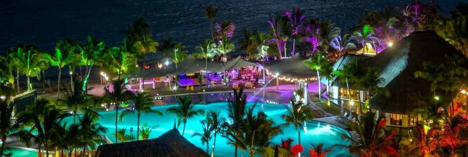 A nighttime tropical resort scene with illuminated pools and colorful lights, surrounded by palm trees near a calm ocean, exuding a relaxing, festive vibe.