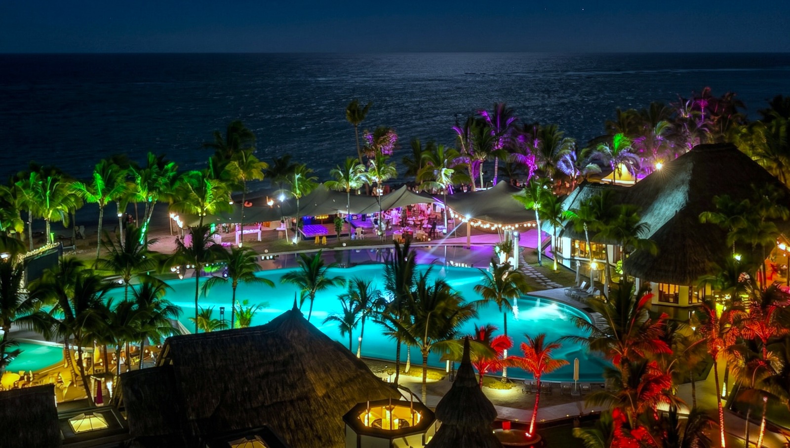 A nighttime tropical resort scene with illuminated pools and colorful lights, surrounded by palm trees near a calm ocean, exuding a relaxing, festive vibe.