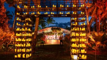 An outdoor evening event featuring a large, rectangular archway constructed from a black metal grid. The grid is filled with hundreds of glowing yellow candles, creating a bright, warm entryway. In the background, a person stands near a dimly lit bar area sheltered by a canopy, surrounded by palm trees illuminated with orange light against a deep blue twilight sky.