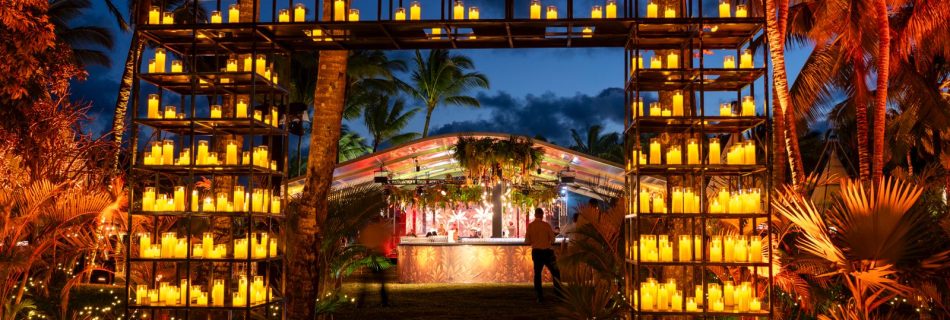 An outdoor evening event featuring a large, rectangular archway constructed from a black metal grid. The grid is filled with hundreds of glowing yellow candles, creating a bright, warm entryway. In the background, a person stands near a dimly lit bar area sheltered by a canopy, surrounded by palm trees illuminated with orange light against a deep blue twilight sky.