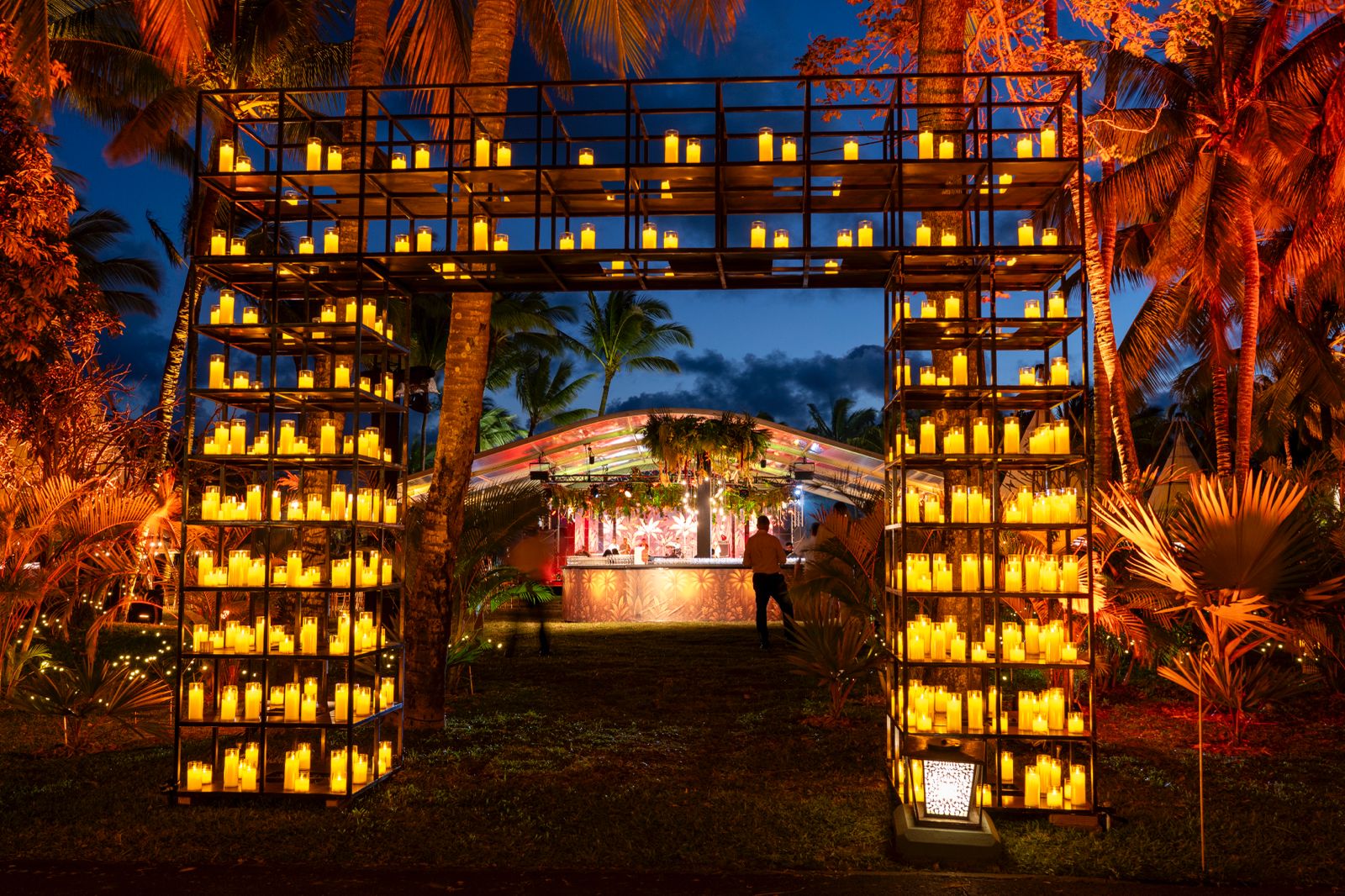 An outdoor evening event featuring a large, rectangular archway constructed from a black metal grid. The grid is filled with hundreds of glowing yellow candles, creating a bright, warm entryway. In the background, a person stands near a dimly lit bar area sheltered by a canopy, surrounded by palm trees illuminated with orange light against a deep blue twilight sky.