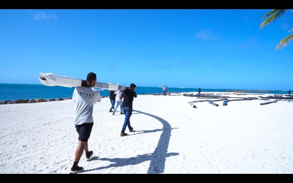 A group of people walking on a white sandy beach carrying long poles, with the blue ocean and clear sky in the background.