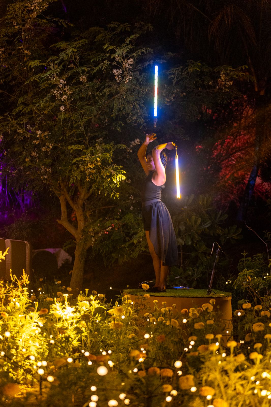 Nighttime performer or dancer holding light-up LED batons on a stage surrounded by warm fairy lights and tropical garden foliage.