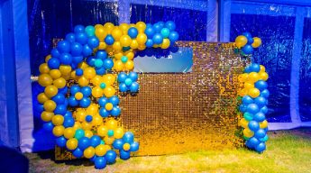Photo booth backdrop with gold sequin shimmer wall and blue and yellow balloon arch for a party or corporate event.