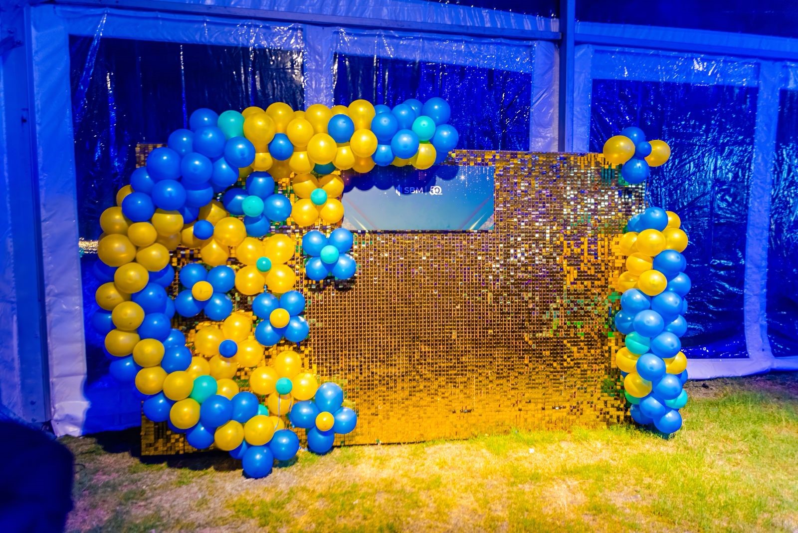 Photo booth backdrop with gold sequin shimmer wall and blue and yellow balloon arch for a party or corporate event.