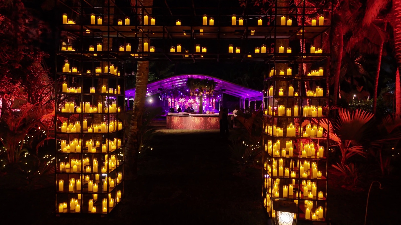 Illuminated garden archway adorned with numerous glowing candles, leading to a vibrant party area with pink and purple lighting, creating a festive ambiance.