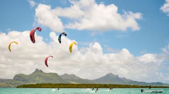 Kite surfers glide over turquoise waters, vibrant kites in the sky. Mountains and lush greenery form a serene backdrop under a partly cloudy sky.