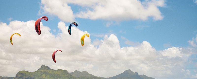 Kite surfers glide over turquoise waters, vibrant kites in the sky. Mountains and lush greenery form a serene backdrop under a partly cloudy sky.