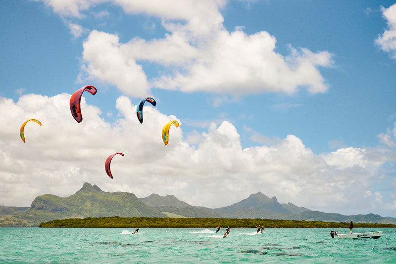 Kite surfers glide over turquoise waters, vibrant kites in the sky. Mountains and lush greenery form a serene backdrop under a partly cloudy sky.
