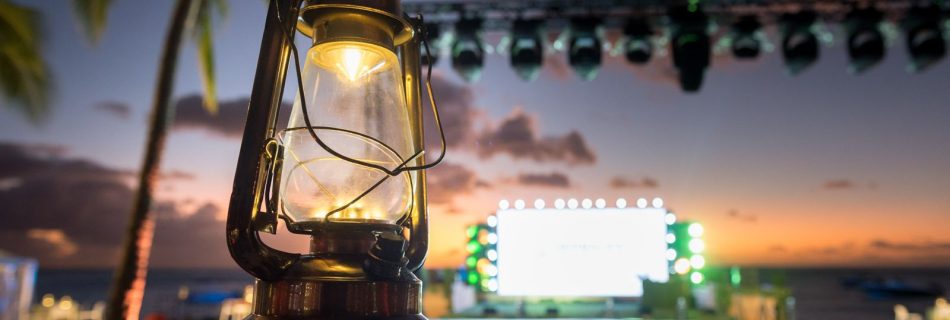 Close-up of a vintage hurricane lantern on a tropical print table during a sunset event with an illuminated stage and palm trees in the background