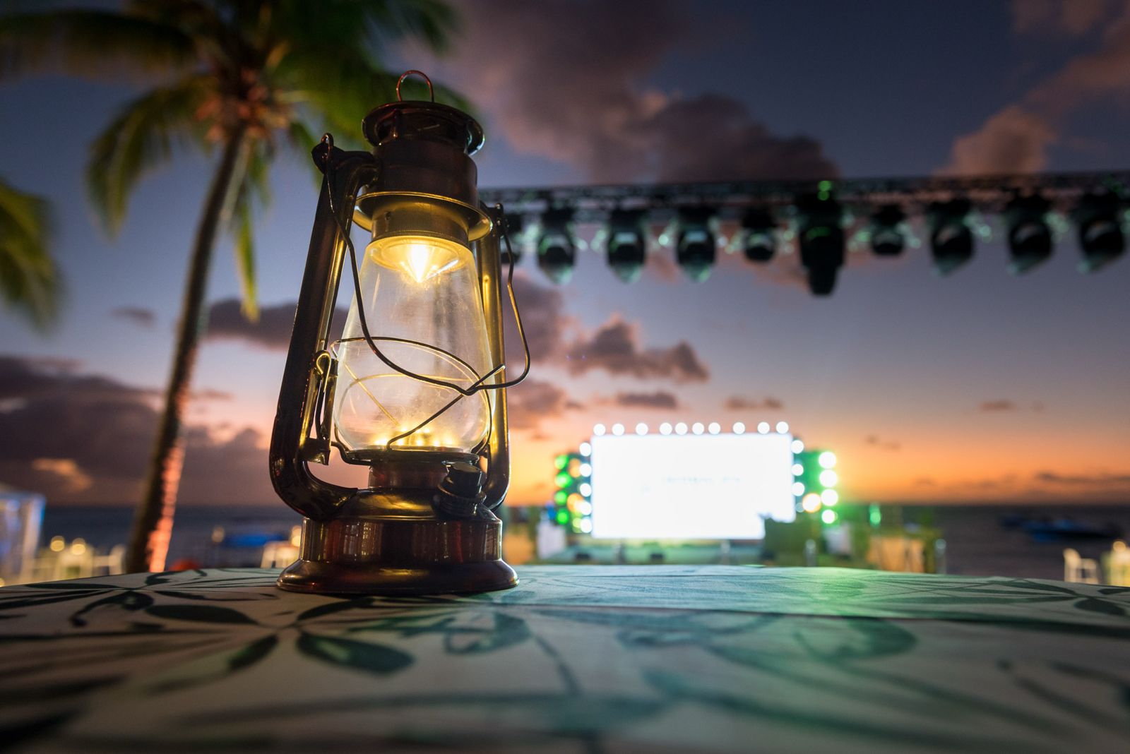 Close-up of a vintage hurricane lantern on a tropical print table during a sunset event with an illuminated stage and palm trees in the background
