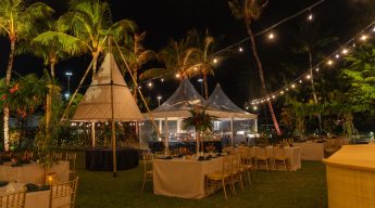Tropical luxury nighttime dinner reception set up on a lawn, featuring a central white tipi tent and smaller clear-span tents, illuminated by warm string lights hung between palm trees over decorated dining tables with gold chairs.