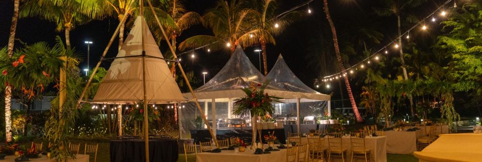 Tropical luxury nighttime dinner reception set up on a lawn, featuring a central white tipi tent and smaller clear-span tents, illuminated by warm string lights hung between palm trees over decorated dining tables with gold chairs.