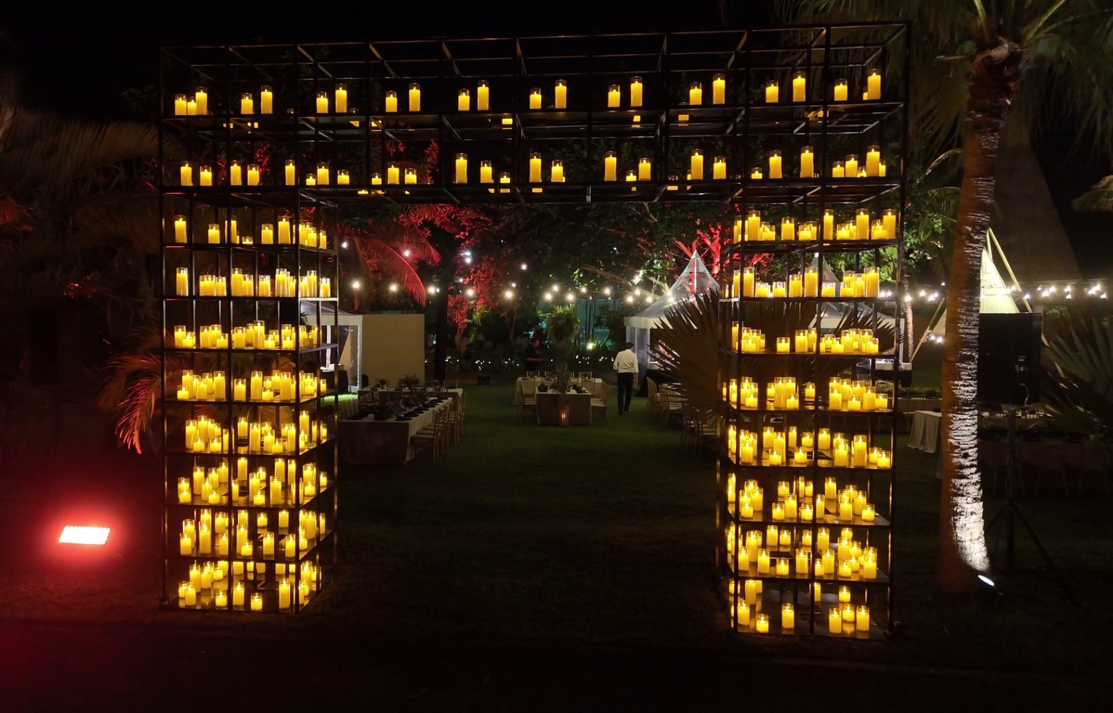 A large metal archway adorned with rows of glowing candles leads to an outdoor evening event. String lights and decorated tables create a warm, inviting atmosphere.