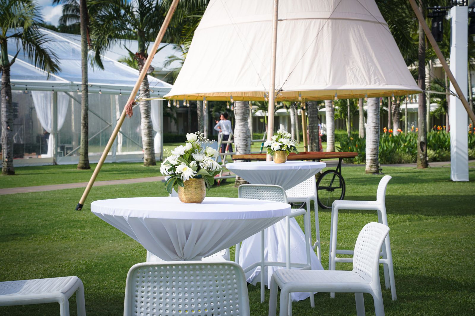 Outdoor cocktail area setup on a green lawn featuring white high-top tables with white floral centerpieces, modern white chairs, and a large white conical or tipi tent structure, surrounded by palm trees.