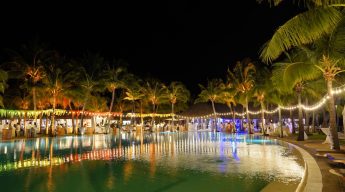 Close-up of luxury poolside event decor featuring golden uplight on palm trees, fringed fabric draping, and slatted wooden cocktail pedestals.