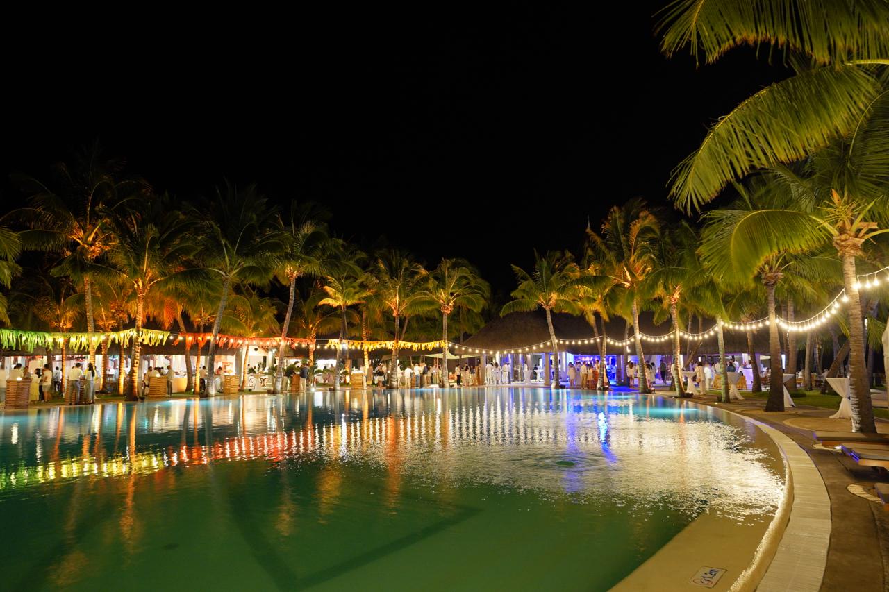 Close-up of luxury poolside event decor featuring golden uplight on palm trees, fringed fabric draping, and slatted wooden cocktail pedestals.