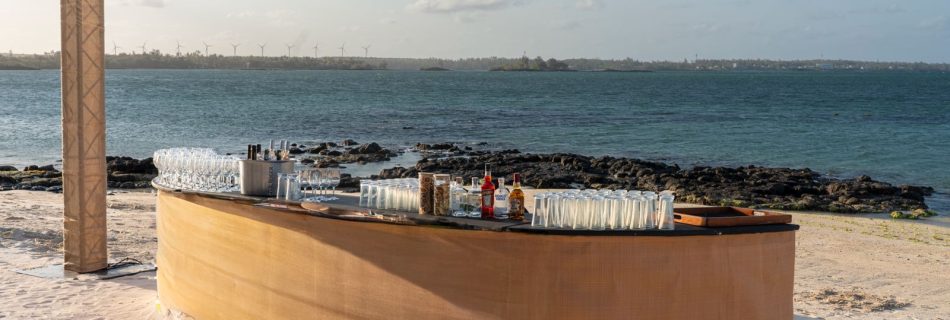 A curved, temporary bar set up on a white sandy beach, fully stocked with glassware and spirits, overlooking a turquoise lagoon.