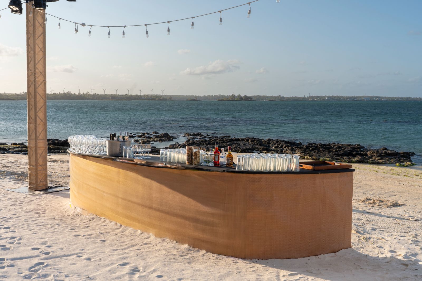 A curved, temporary bar set up on a white sandy beach, fully stocked with glassware and spirits, overlooking a turquoise lagoon.