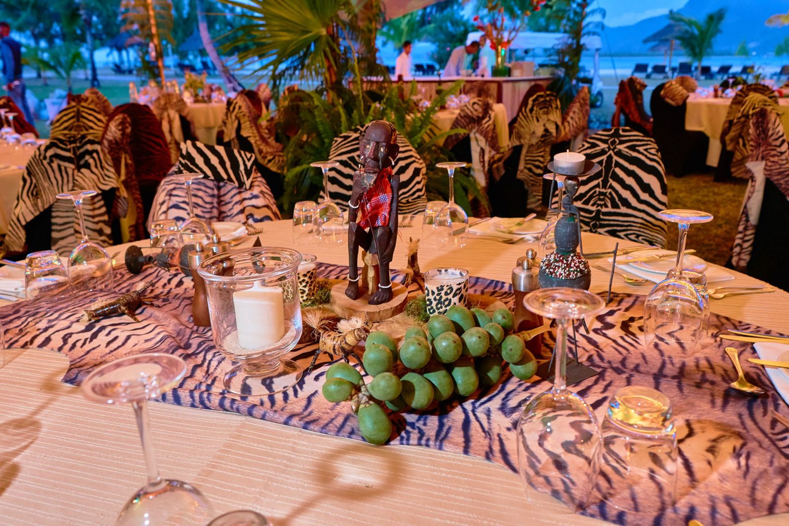 An outdoor dinner table setup with an African safari theme, featuring a tiger print table runner and chair covers, a wooden figurine centerpiece, candles in glass holders, and green fruit or vegetables arranged on the runner.