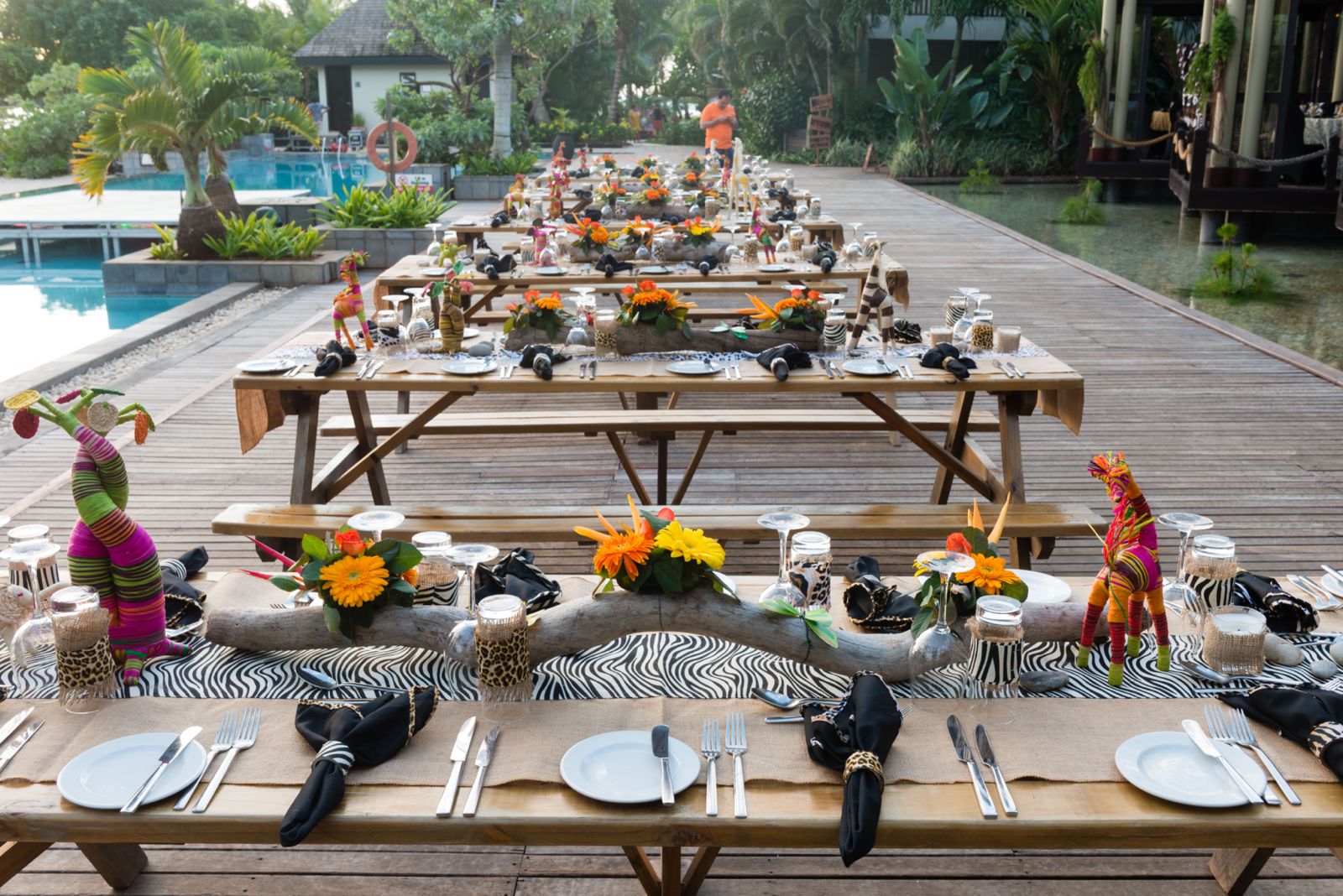 A long series of picnic tables set up outdoors on a wooden deck next to a pool, decorated with an African safari theme, featuring zebra print runners, driftwood centerpieces, bright orange and yellow flowers, and colorful woven animal figurines.