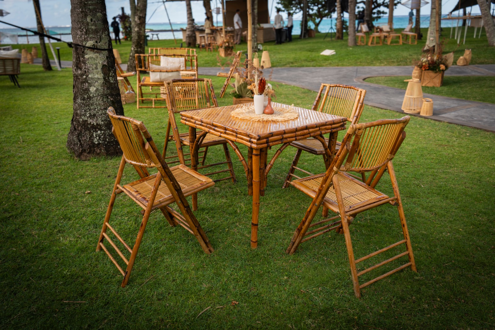 Bamboo dining set with four chairs on a lush green lawn, under tall trees. Decorated table with a vase; relaxed, outdoor setting near the ocean.
