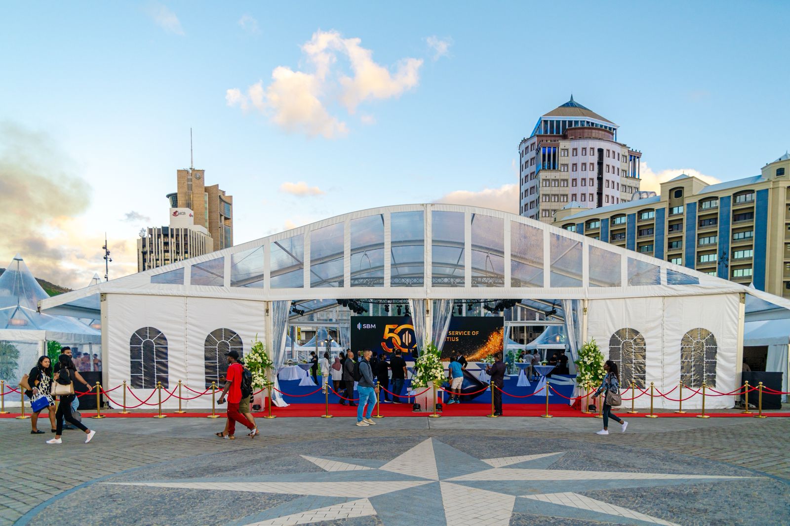 A large white event tent with a clear roof structure, set up in an urban square with a star pattern on the ground and modern high-rise buildings in the background, showing people entering the tent on a red carpet.