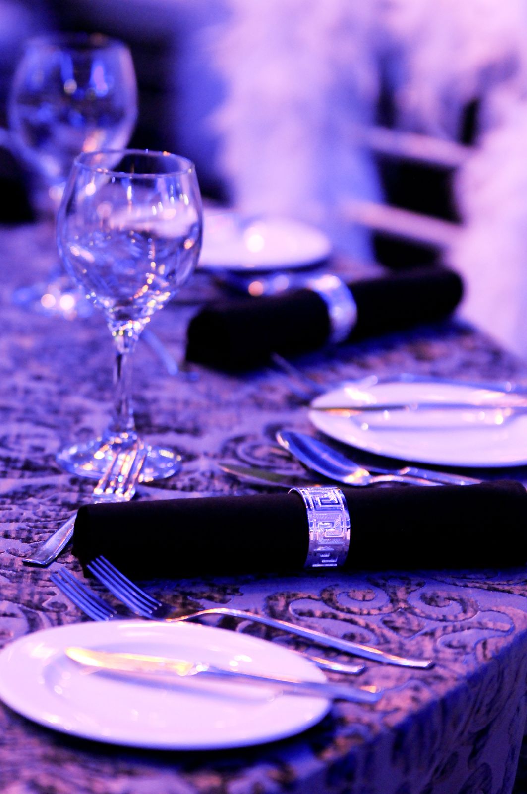 A close-up view of an elegant dinner table setting featuring white plates, silverware, a black cloth napkin held by a silver, patterned napkin ring, and a wine glass, all resting on a patterned tablecloth under bright purple lighting.