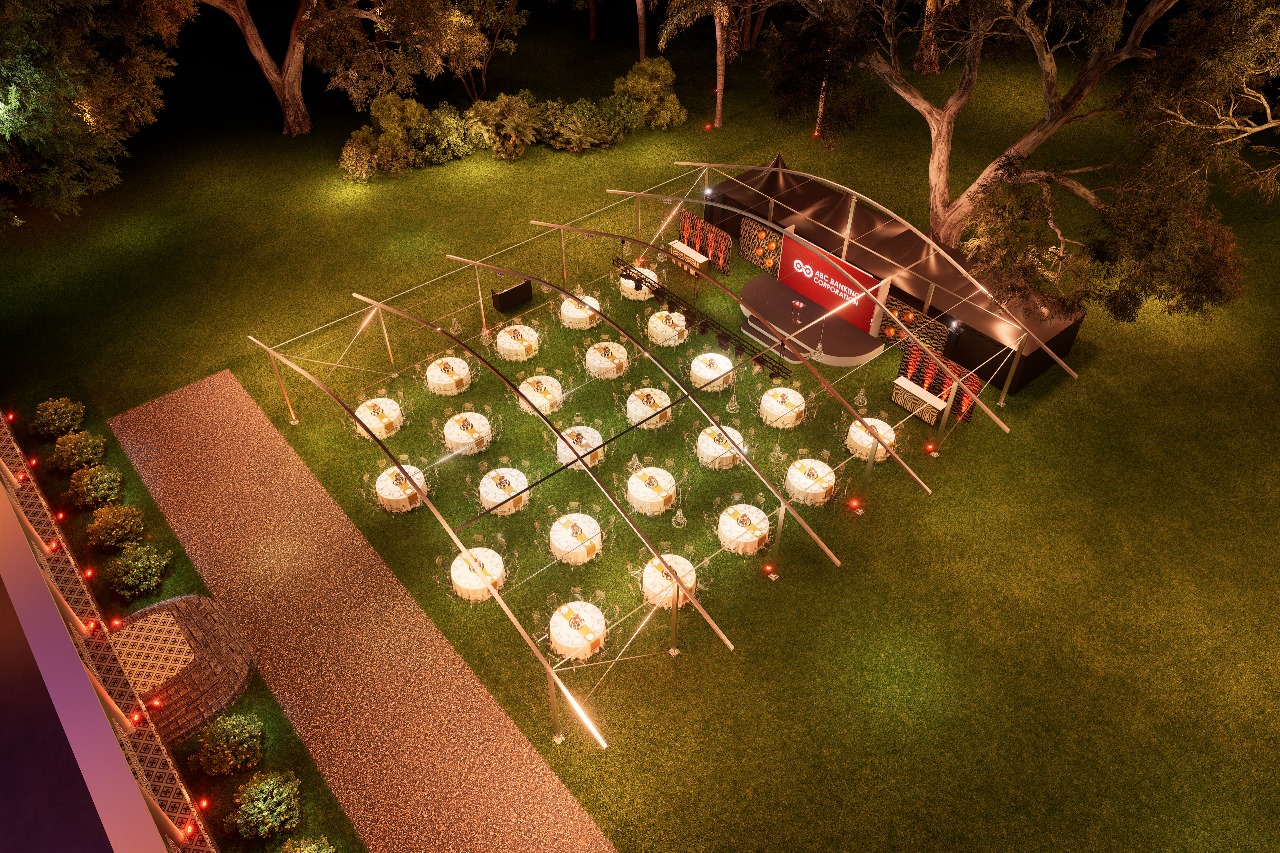 An overhead nighttime view of a sophisticated, temporary event venue set up on a beach. A large, open-air tent covers multiple round dinner tables, each lit by a warm, glowing centerpiece, facing a stage with large screens. To the right of the tent is a wooden deck area with a circular bar or lounge and several large, illuminated, teepee-like cabanas, all surrounded by palm trees and dark sand.