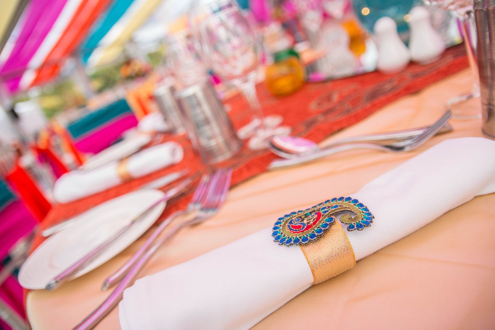 A close-up of a festive table setting featuring a white cloth napkin wrapped with a gold band and secured by a brightly colored, enamel paisley-shaped napkin ring, with colorful draped fabrics visible in the background.