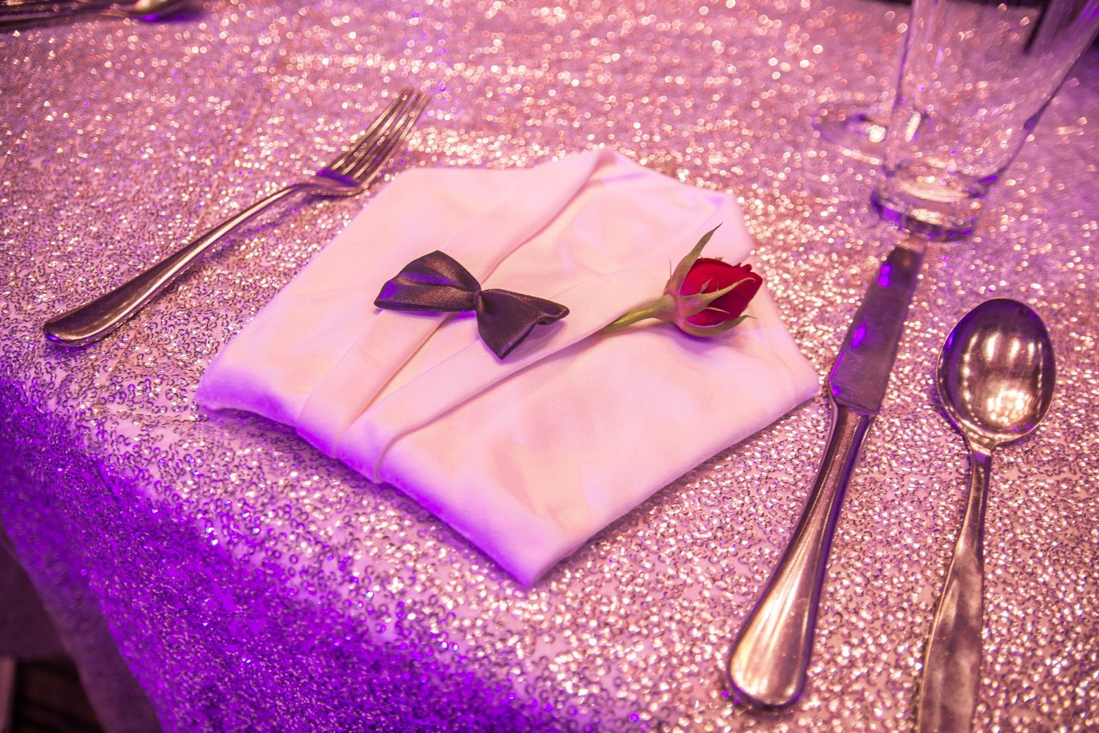A formal dinner place setting featuring a white, folded cloth napkin shaped like a dinner jacket, adorned with a miniature black bowtie and a single red rosebud, surrounded by silverware on a shimmering, sequined pink and purple tablecloth.