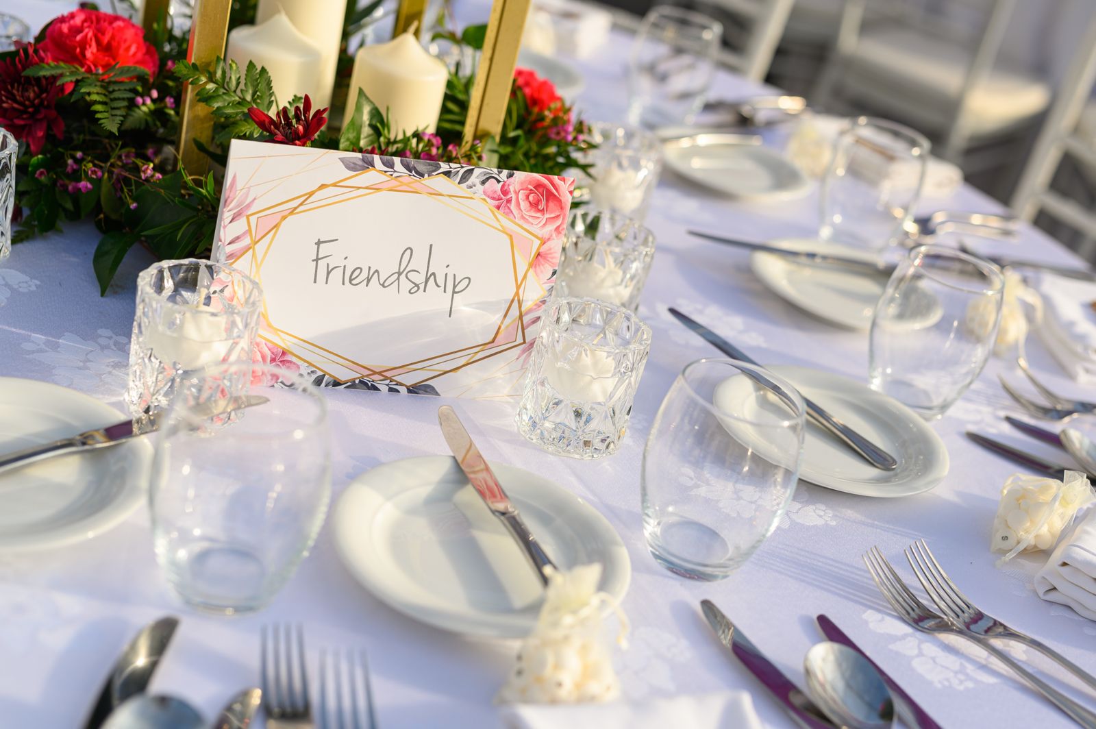 A wedding or formal dining table set with white plates and linens, centered around a table card featuring the word "Friendship" inside a gold geometric frame with pink and red floral accents, surrounded by candles and a floral centerpiece.