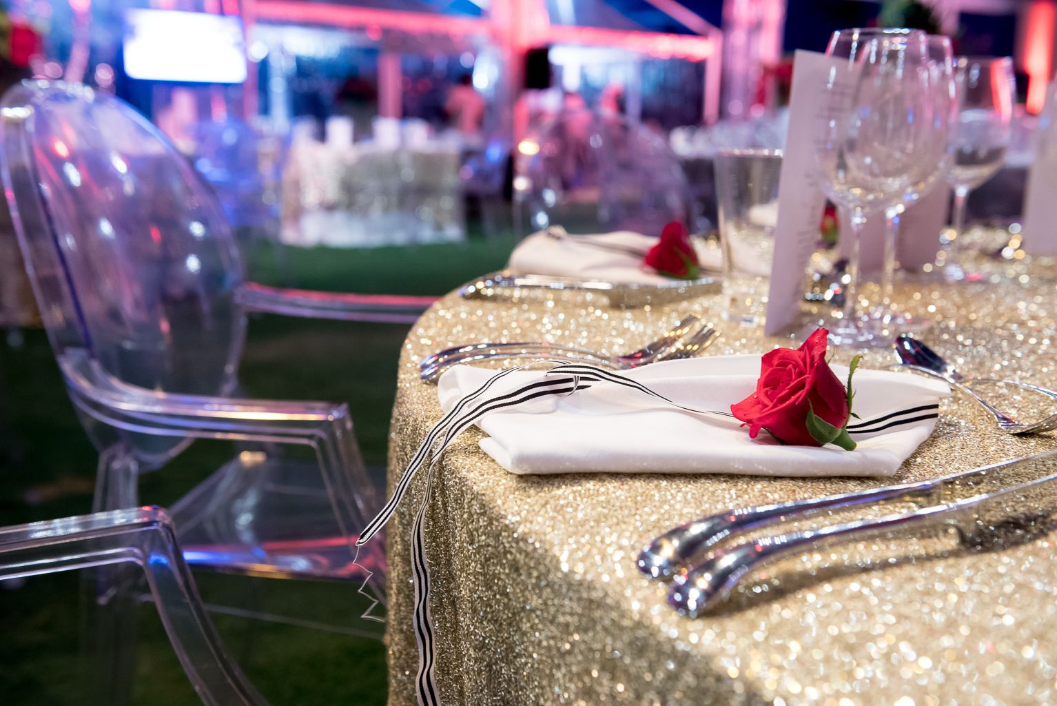 An elegant dinner table setup at an event, showing a clear acrylic ghost chair next to a round table covered in a shimmering gold sequined tablecloth, with a place setting featuring a white napkin, black and white striped ribbon, and a bright red rose.