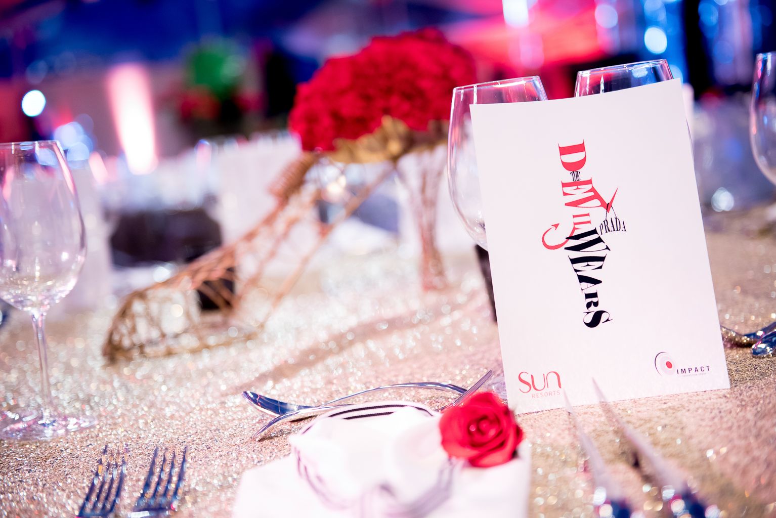 A glamorous table setting featuring a menu card with the title "Devil Wears Prada" in striking red and black text, placed next to wine glasses on a sparkling gold sequined tablecloth, and decorated with a single red rose on a white napkin.