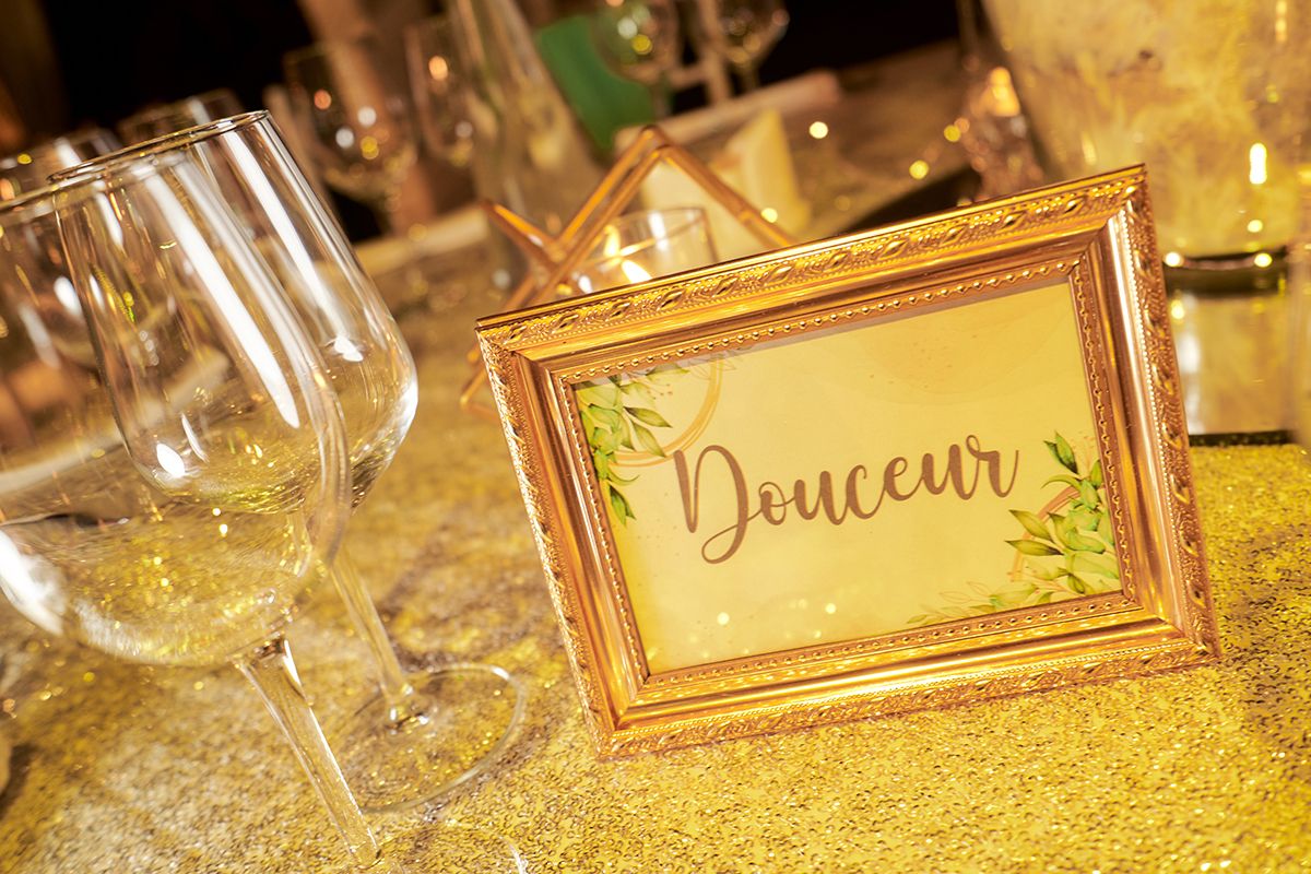 A close-up of a golden-framed table sign with the word "Douceur" (Sweetness) written in cursive on a yellow background, surrounded by sparkling gold tablecloth and clear wine glasses.