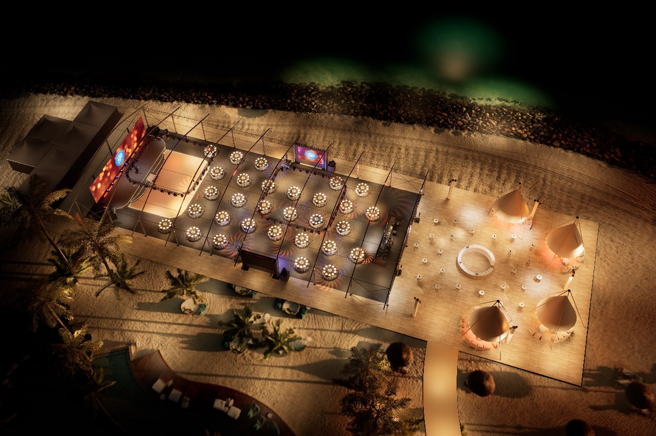 An overhead view of an outdoor evening event setup on a lush green lawn, surrounded by trees. Twenty-four round dinner tables with glowing centerpieces are arranged in rows under a simple wire frame structure. A stage with a curved canopy and a backdrop featuring a red and black logo (possibly '60 Years') is positioned at the back, next to a large tree.