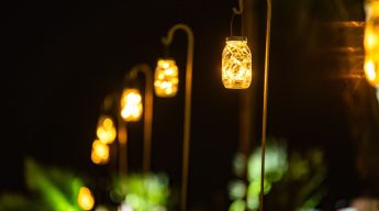 Close-up of glowing mason jar lanterns filled with fairy lights, hanging from curved shepherd's hooks, creating a warm, inviting pathway light effect at a nighttime outdoor event.