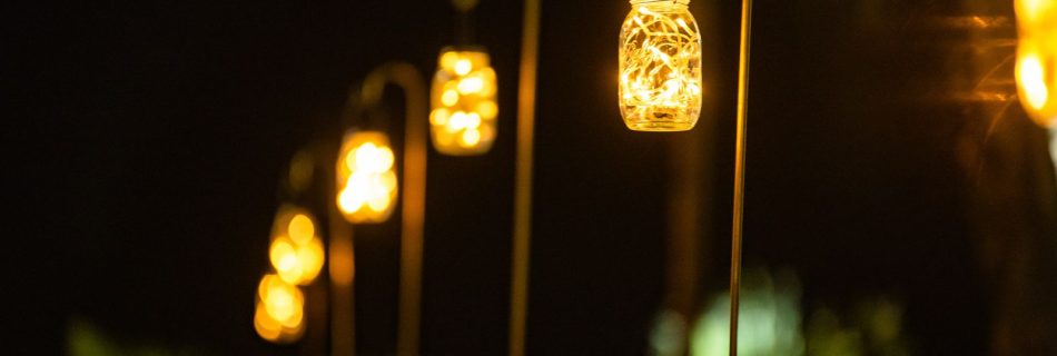 Close-up of glowing mason jar lanterns filled with fairy lights, hanging from curved shepherd's hooks, creating a warm, inviting pathway light effect at a nighttime outdoor event.
