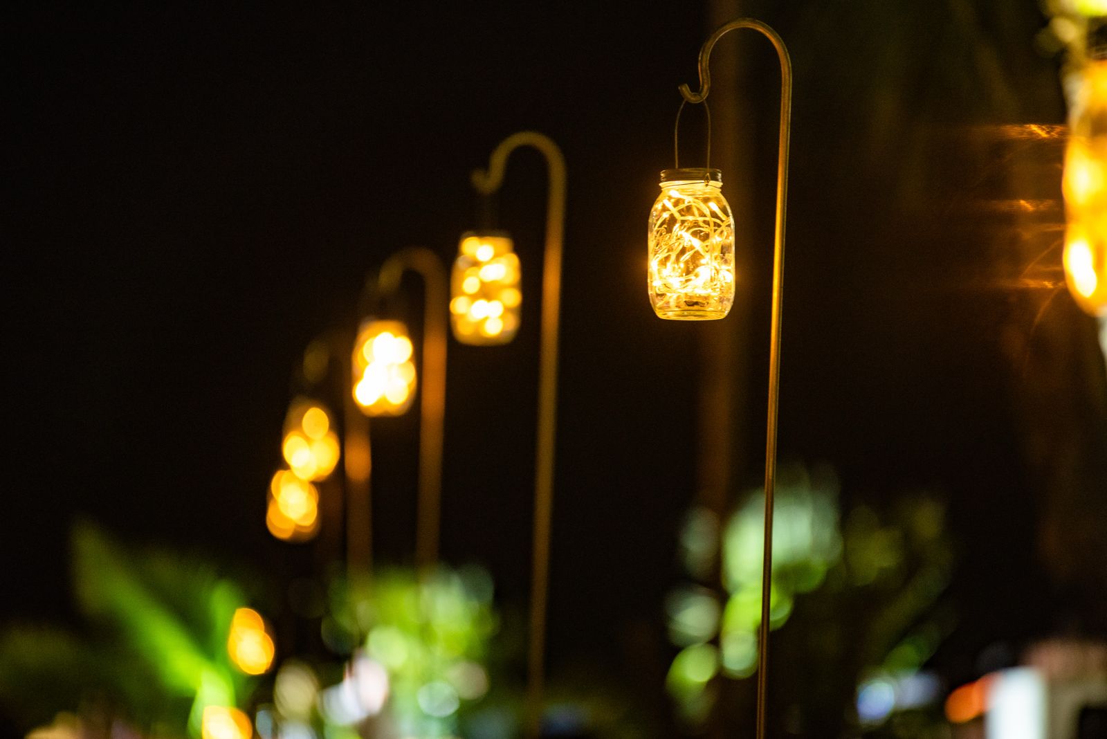 Close-up of glowing mason jar lanterns filled with fairy lights, hanging from curved shepherd's hooks, creating a warm, inviting pathway light effect at a nighttime outdoor event.