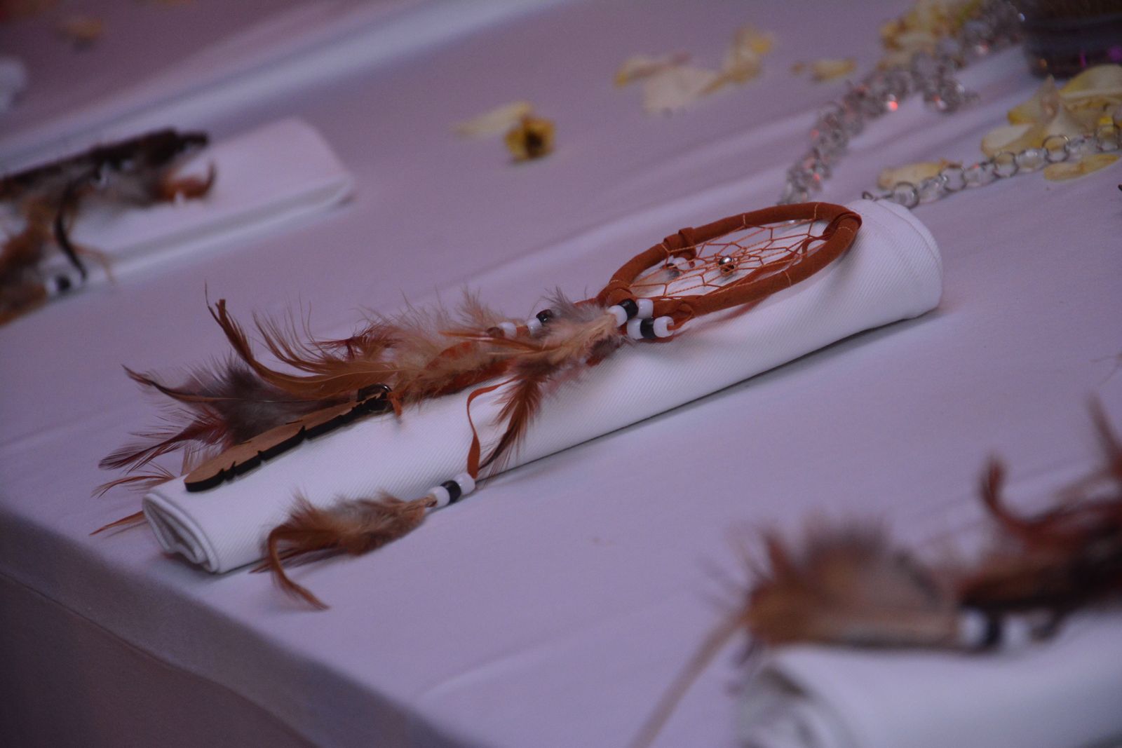 A small, miniature dreamcatcher with brown feathers and beads, resting on a neatly rolled white cloth napkin on a white or light purple tablecloth, used as a decorative place setting.
