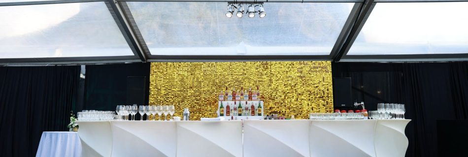 Contemporary white curved bar setup with bottles, glassware, and a striking gold sequin shimmer wall backdrop inside a clear marquee.
