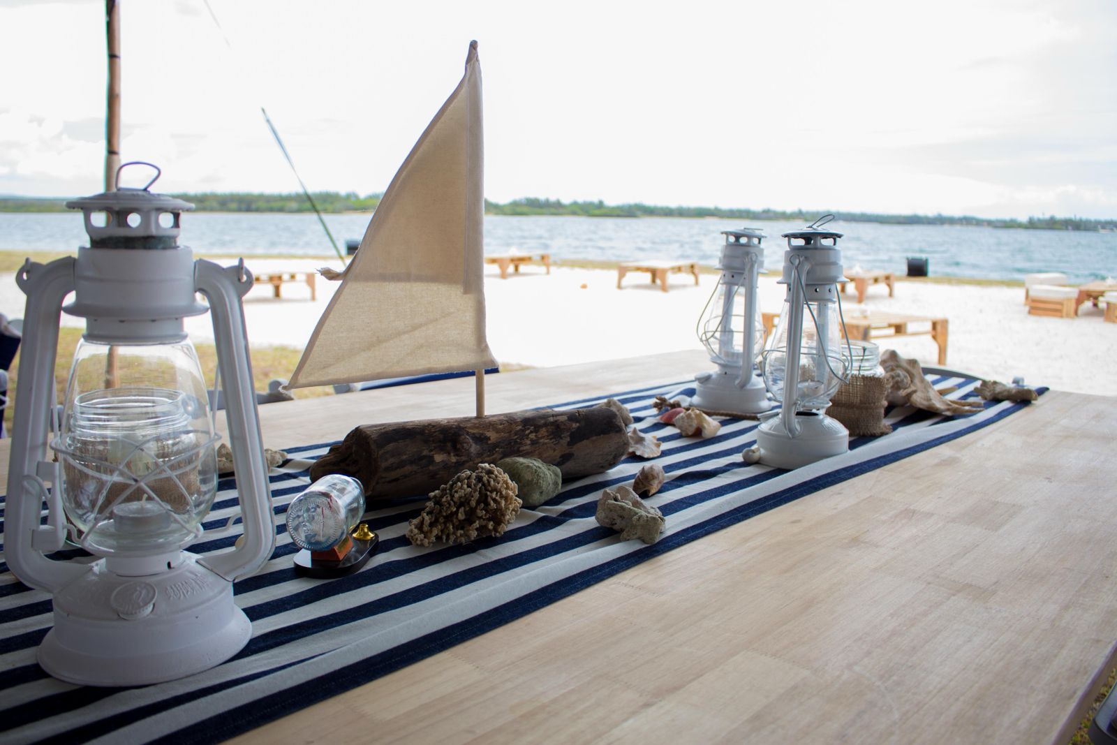 A nautical-themed table decoration featuring two white kerosene lanterns, a small sailboat model with a tan sail, driftwood, seashells, and coral, all arranged on a blue and white striped table runner, with a sandy beach and water visible in the background.