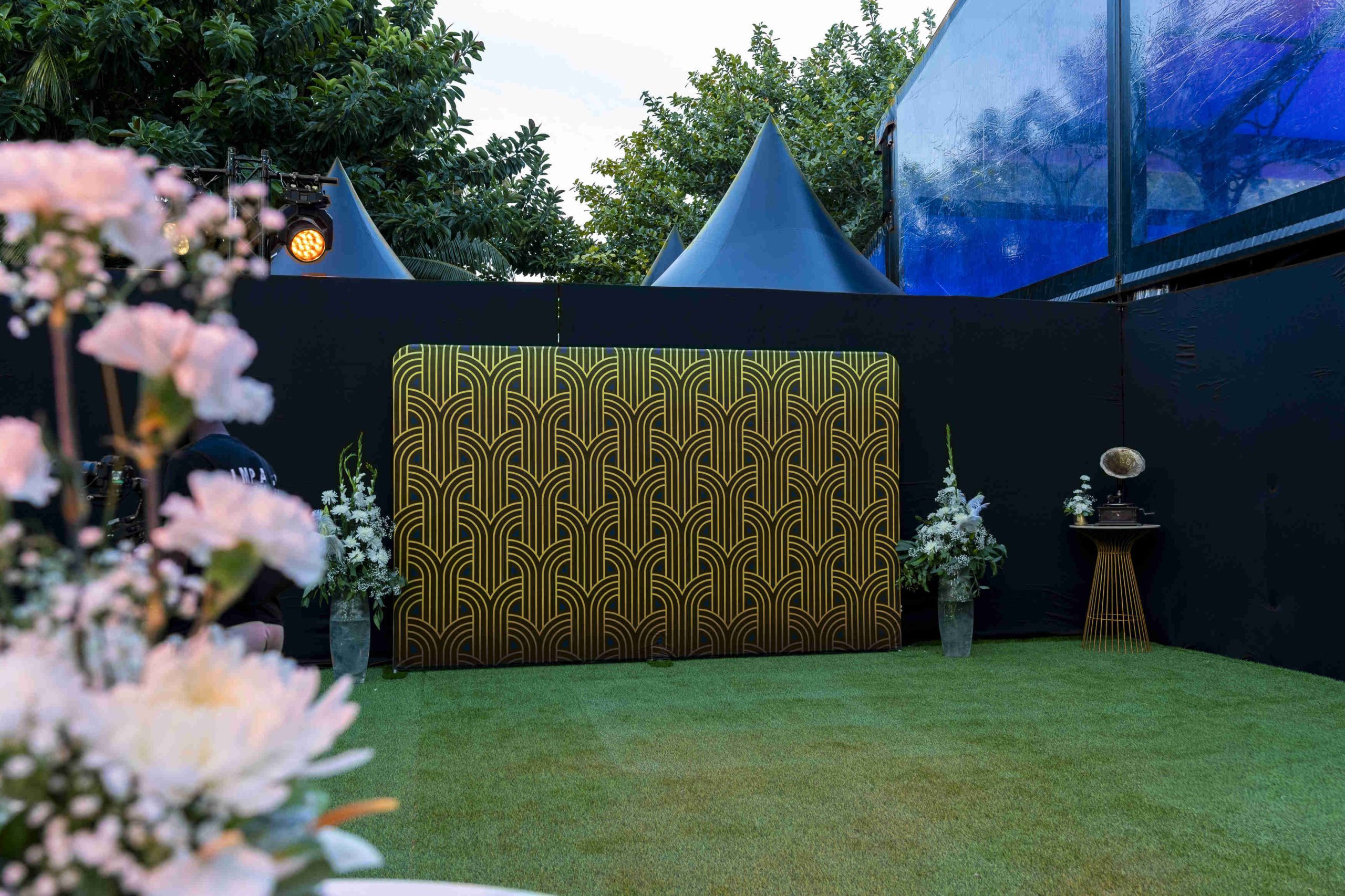 Outdoor event setup with art deco-patterned screen, surrounded by vases of white flowers on grass. Dark tent tops and trees in the background.