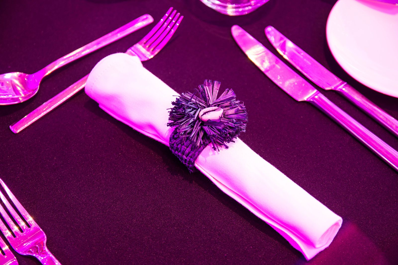 A close-up of a place setting featuring a white rolled napkin secured by a dark, textured napkin ring with a seashell centerpiece, set on a dark tablecloth under intense magenta or pink lighting that illuminates the silverware.