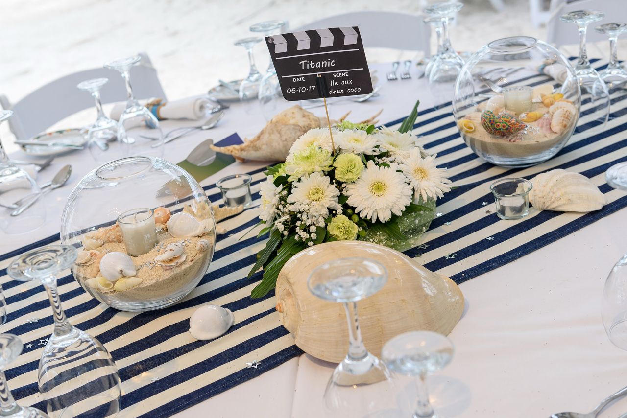 A nautical or beach-themed table centerpiece featuring a black movie clapperboard sign reading "Titanic" and "Ile aux deux cocos," placed next to a bouquet of white flowers, surrounded by glass bowls filled with sand and seashells, and large conch shells, all on a blue and white striped table runner.