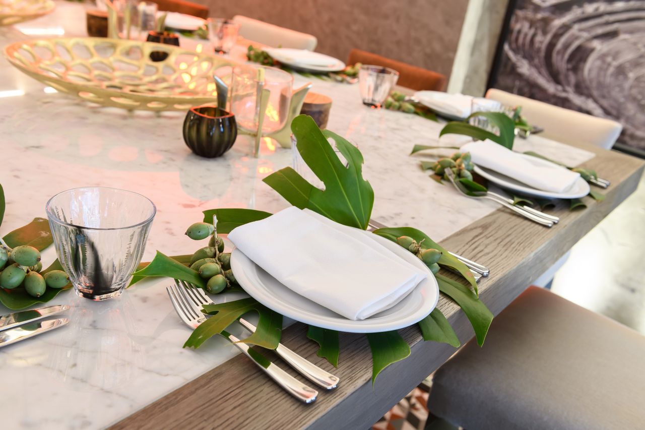 A contemporary table setting on a marble table with wooden accents, featuring a white plate and napkin, decorated with large green tropical leaves and clusters of green berries, and includes modern glassware and silverware.