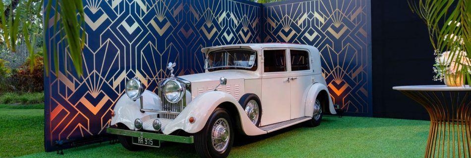 Outdoor event crew setting up black flooring in front of a white vintage car and a black and gold Art Deco backdrop under tropical palm trees.