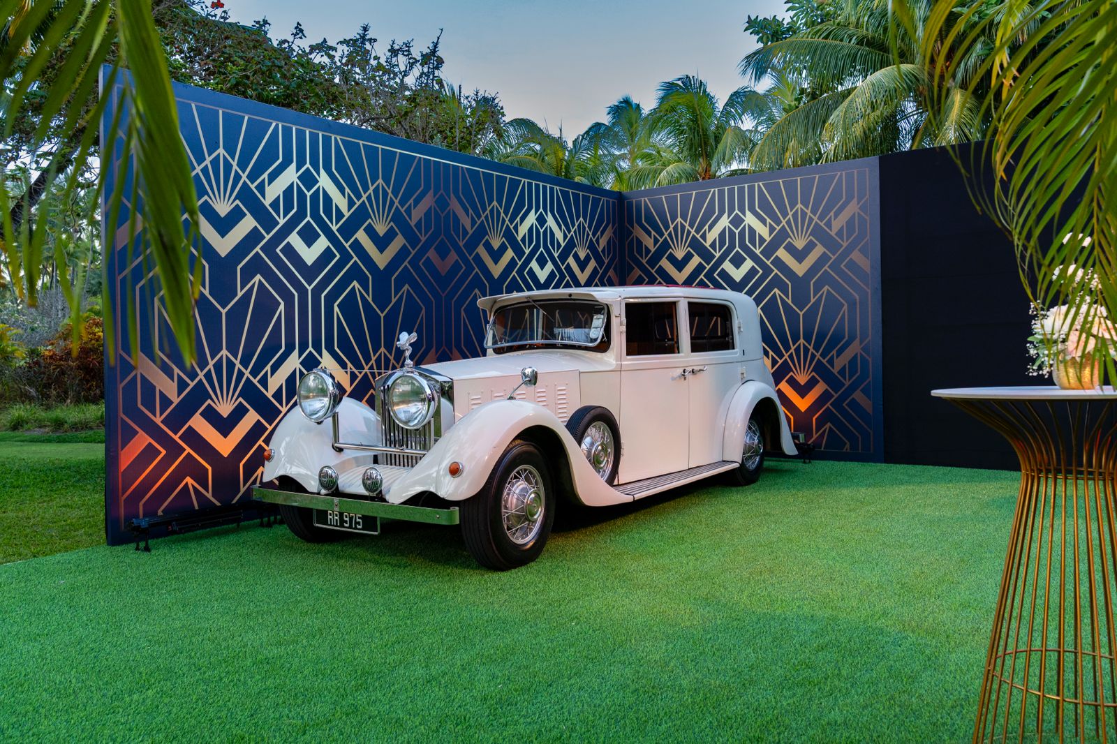 Outdoor event crew setting up black flooring in front of a white vintage car and a black and gold Art Deco backdrop under tropical palm trees.