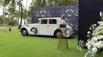 Vintage white car parked on lush green grass beside a geometric-patterned wall. A decorative table holds a record player, with flowers in the foreground.