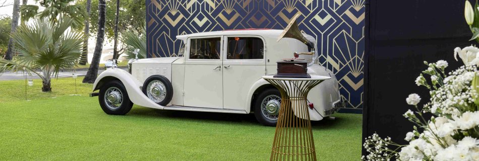 Vintage white car parked on lush green grass beside a geometric-patterned wall. A decorative table holds a record player, with flowers in the foreground.
